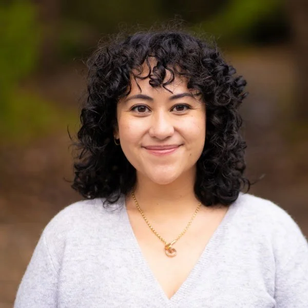 A young woman with dark curly hair, wearing a light gray sweater and a gold necklace, smiling outdoors with blurred greenery in the background.