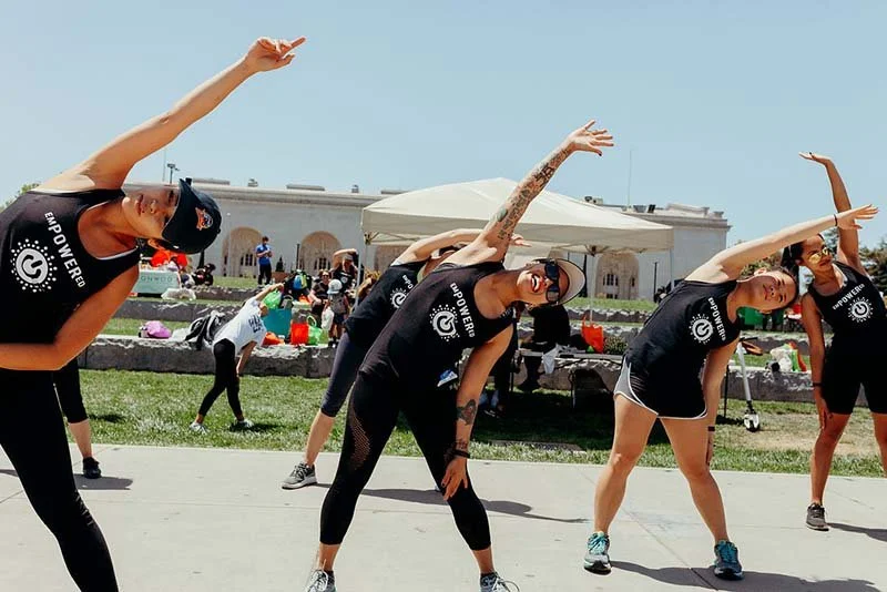 Group of women performing a side stretch exercise outdoors, wearing matching black tank tops with a logo, in front of a white tent at a public event.