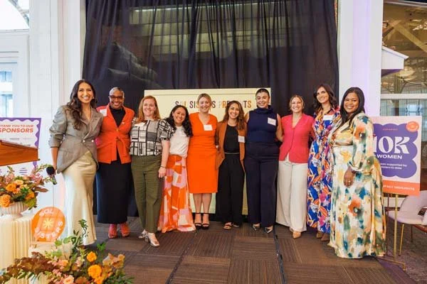 Group of ten women standing together indoors at an event, smiling for the photo, with flowers and signage in the background.