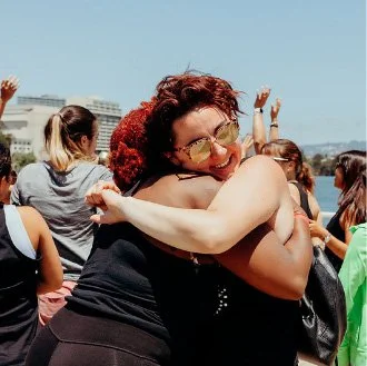 A woman with curly red hair and glasses hugging another woman outdoors, surrounded by several people on a sunny day.