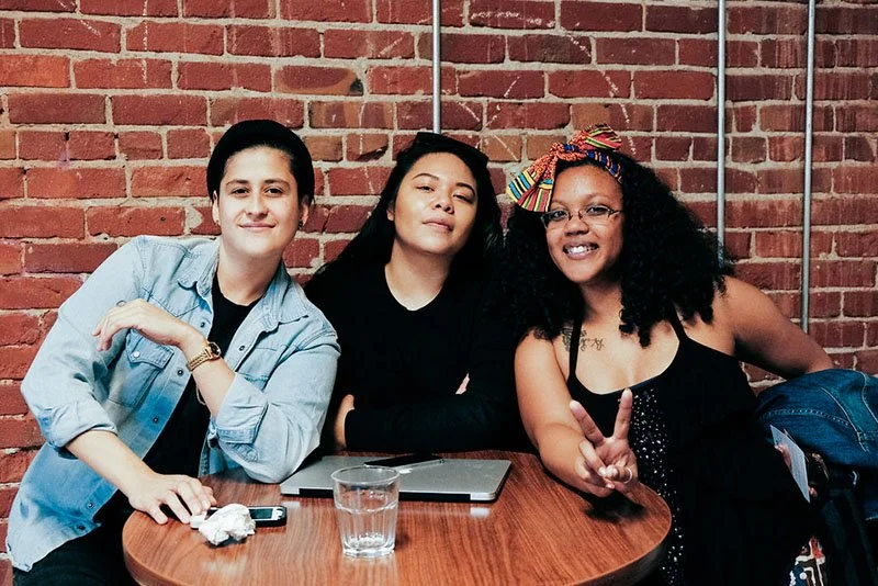 Three women sitting at a round table against a brick wall, smiling and making peace signs. One wears a denim jacket, one a black top, and the third an afro hairstyle with a colorful headband and glasses.