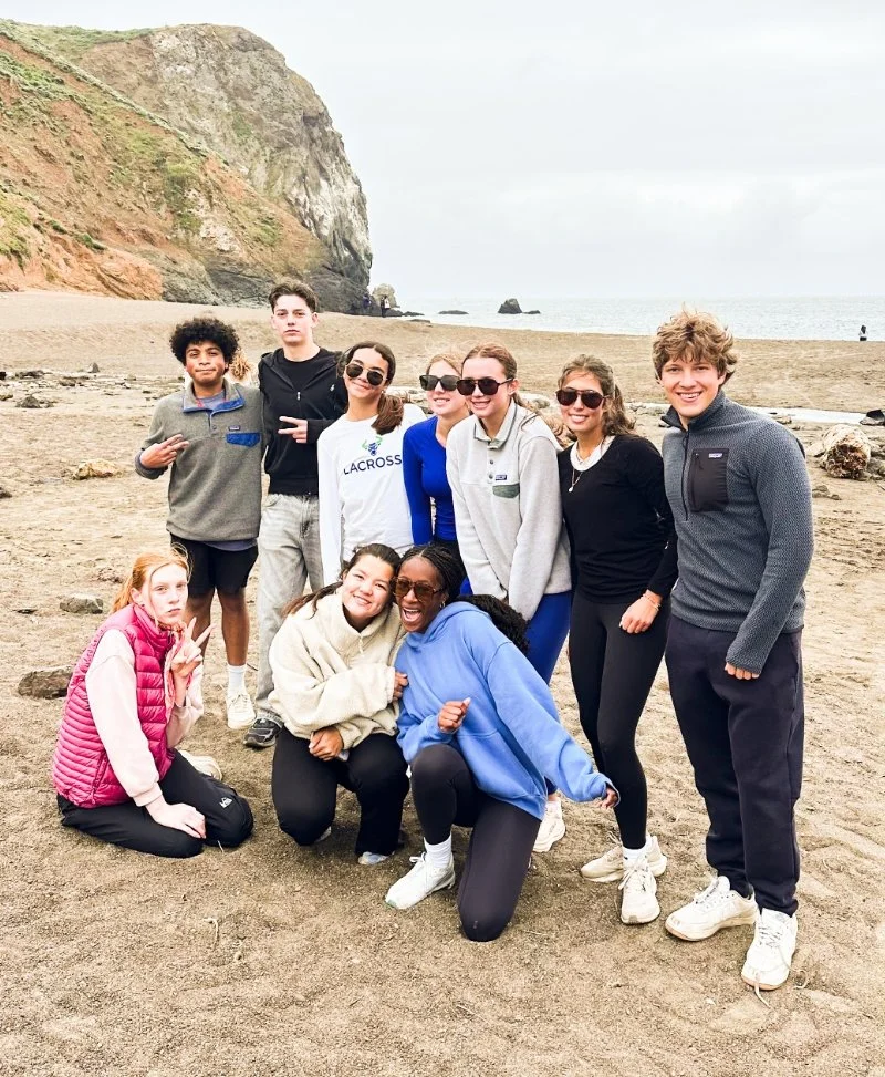 A group of ten teenagers, standing and kneeling, on a sandy beach near a rocky cliff, smiling and posing for the photo on an overcast day.