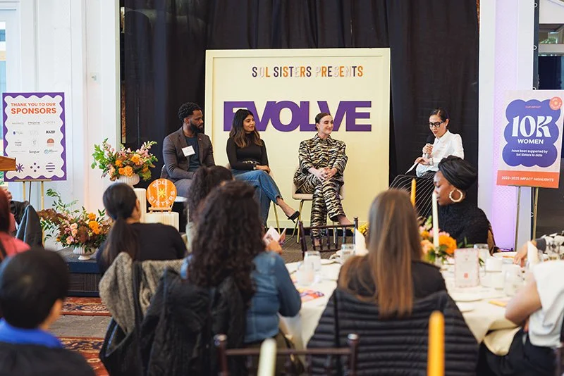 Women speaking on a panel at the Sol Sisters event titled 'EVOLVE,' with an audience of women seated at tables in a conference room decorated with flowers and banners.