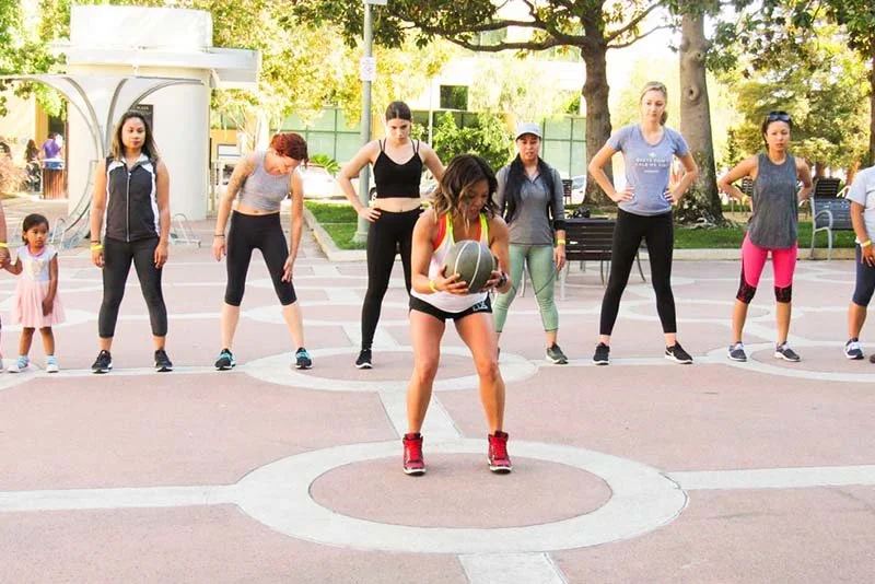 A woman is holding a basketball and performing a workout or exercise in an outdoor gym area, with several women standing behind her in a line, some with hands on hips or arms crossed, in a park with trees and sunlight.