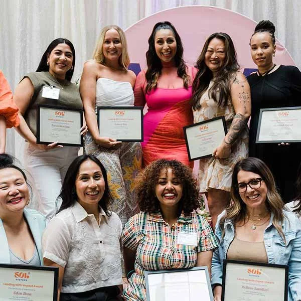 Group of ten women at an awards ceremony, with seven women standing in the back row holding certificates, and three women seated in the front, also holding certificates, in front of a pink backdrop.