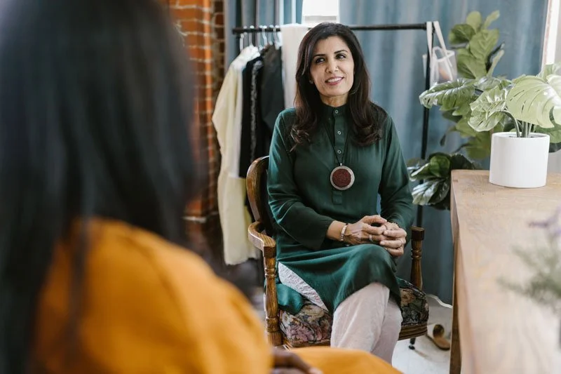 A woman in a green dress sitting in a wooden chair, smiling during a conversation, with a woman in an orange top blurred in the foreground. The setting appears to be a cozy indoor space with plants and clothing racks.