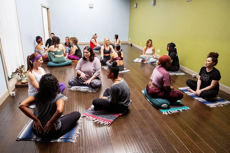Women participating in a yoga or meditation class in a studio with wooden floors and colorful walls, sitting on mats and engaging in conversation or meditative activities.