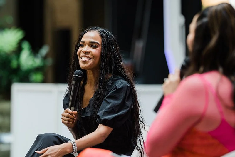 A woman with long braided hair smiling and holding a microphone during a conversation or interview, with another woman in pink sitting nearby.