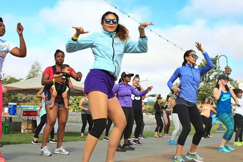 Group of people, mostly women, participating in outdoor fitness dance class in a park on a sunny day.
