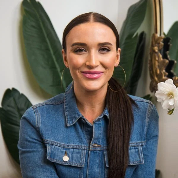 A woman with long brunette hair, wearing a denim jacket and large hoop earrings, smiling in front of green tropical leaves and a decorative wall mirror.