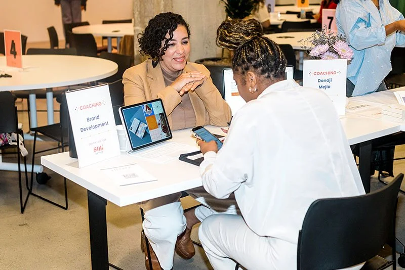 Two women engaged in a coaching session at a table with devices and signs indicating coaching topics.