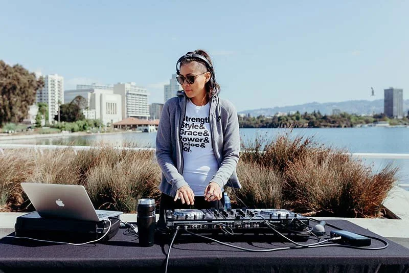 A woman DJing outdoors by a body of water with city buildings in the background.