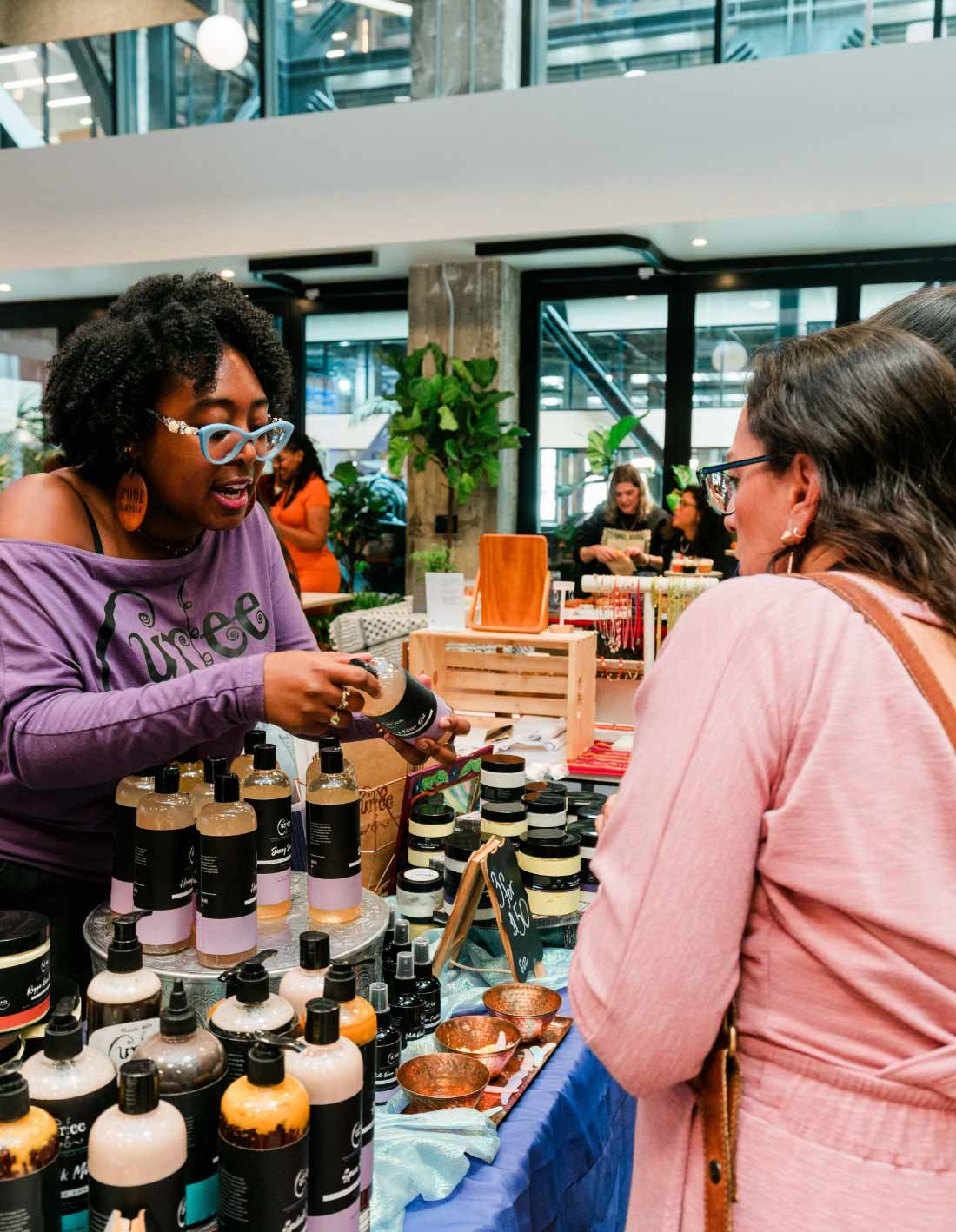 A woman at a market stall showing skincare or beauty products to another woman in a pink jacket, with various bottles and jars displayed on the table.