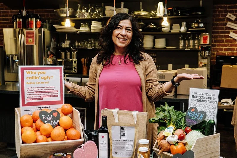 Woman smiling at a farmers market stand with oranges, vegetables, and informational signs.