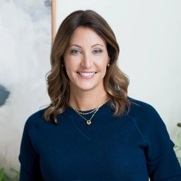 A woman with shoulder-length brunette hair, wearing a dark blue sweater and layered necklaces, smiling at the camera indoors.
