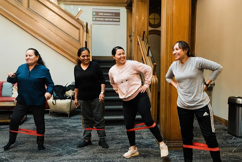 Four women standing in a room, laughing and smiling while participating in a team-building activity using red resistance bands around their legs.