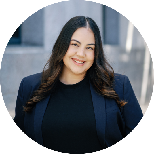 Smiling woman with long dark hair wearing a black shirt and navy blazer, standing outdoors in front of a building.