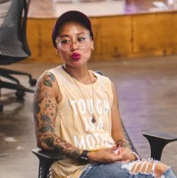 Young woman with tattoos, glasses, and a cap sitting on a chair with wooden flooring and furniture in the background