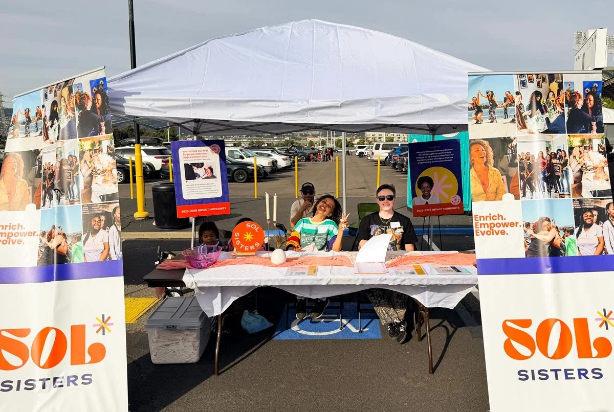 A booth at an outdoor event with SOL Sisters banners, three smiling women sitting behind a table covered with flyers and materials, and other people in the background in a parking lot.