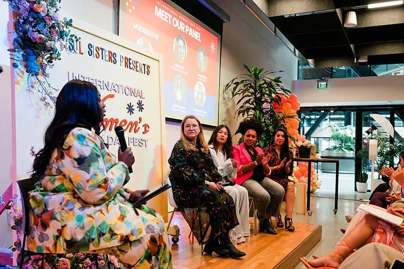 Women participating in a panel discussion at an event themed 'Women's Day Fest' with a floral backdrop and a large screen displaying panel details.
