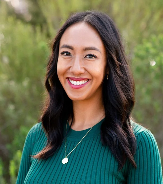 photo of Kate Hermosura smiling at the camera standing outdoors with greenery in the background. she's wearing a teal sweater and has long dark hair and is wearing red lipstick.