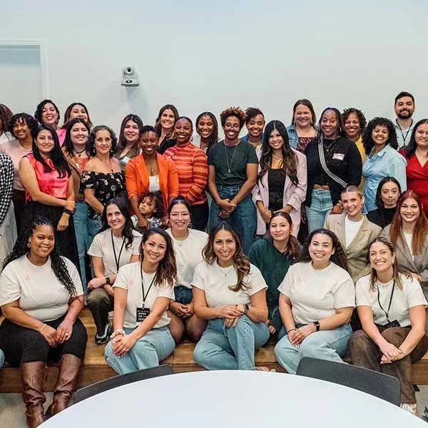 Group photo of diverse women and a young girl in a room with white walls and a wooden platform, smiling at the camera.