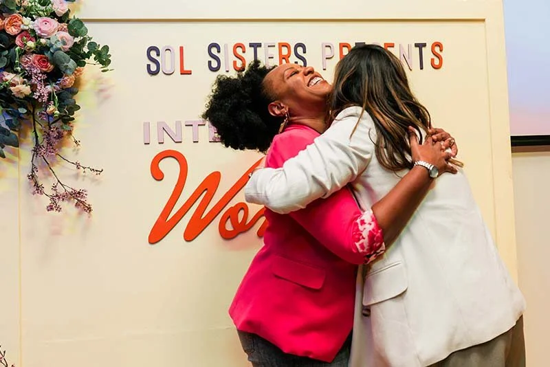 Two women hugging, one wearing a pink blazer and the other in a white blazer, at an event with a backdrop that reads 'SOL SISTERS PRENT' and 'International Women' with floral decorations.