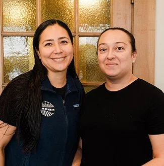 Two women standing indoors, smiling, with a window behind them.
