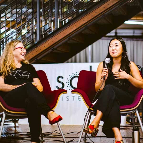 Two women sitting on stage in conversation, one holding a microphone, in front of a backdrop with partial text, against a modern interior with stairs.