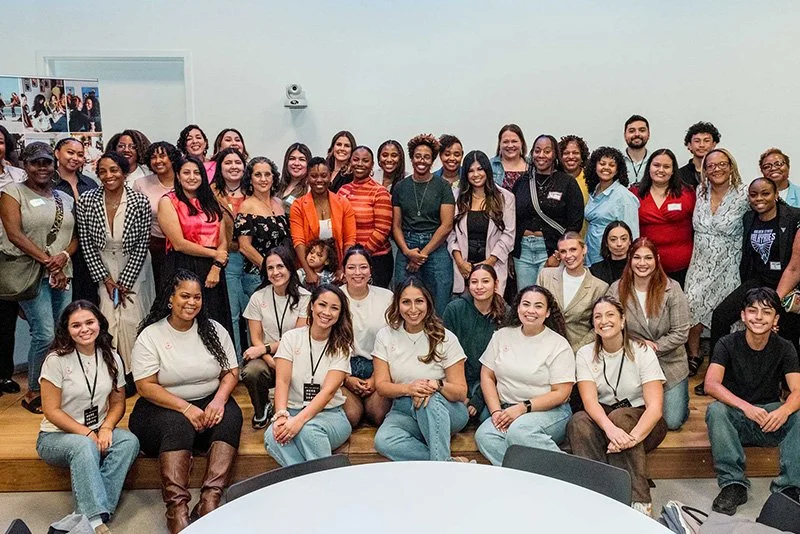 Group of diverse young adults gathered in a room for a photo, some standing and some sitting, with a white wall and poster in the background.