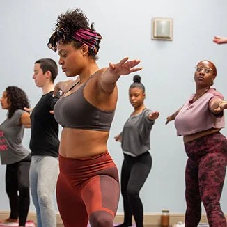 A diverse group of women participating in a yoga or meditation class, standing with arms extended forward and eyes closed in a pose.