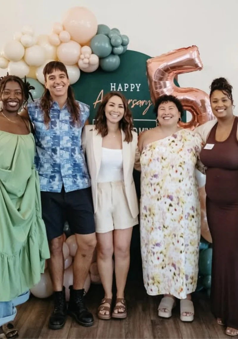 A group of five diverse women standing together, smiling at a celebration event with balloons and a large rose gold number 5 balloon in the background.