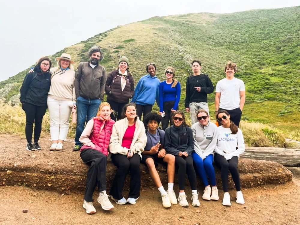 Group of people posing outdoors on a hillside with green grass and rocks, some sitting and some standing, dressed in casual outdoor clothing.
