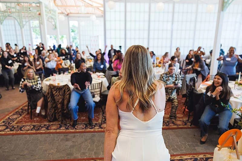 A woman with long blonde hair in a white dress addresses a diverse group of women at a large indoor banquet or conference. Attendees are seated at tables, some smiling and some raising their hands, in a well-lit room with large windows and a high ceiling.