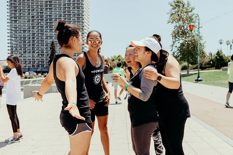 Group of women in athletic clothing chatting and smiling outdoors in a city park on a sunny day.