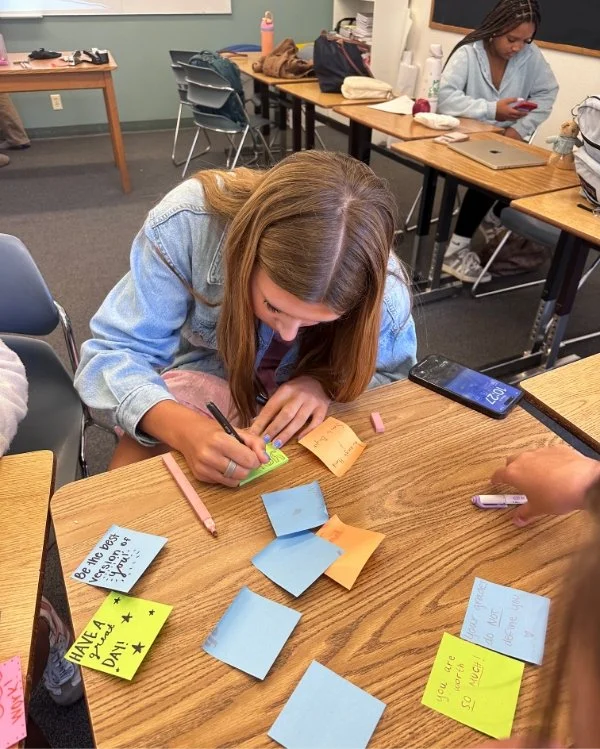 A young woman writing on sticky notes at a classroom table with other students, some using their phones, and classroom supplies visible.