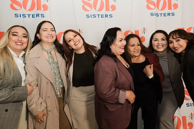 Group of seven women smiling and posing together at an event with a Sol Sisters backdrop.