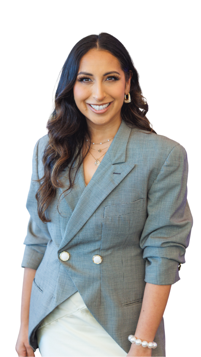 A smiling woman with long dark wavy hair, wearing a gray blazer, pearl jewelry, and light-colored skirt.
