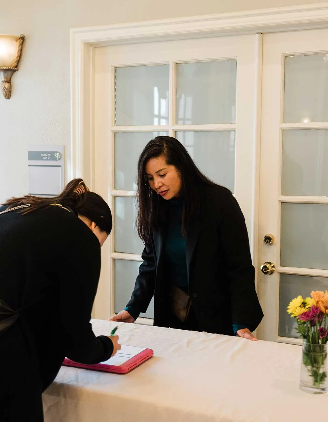 Two women are standing at a table, one is writing on a clipboard, and the other is looking down, leaning on the table. There is a vase with yellow and pink flowers on the right side of the table. They are inside a room with a white door with glass panes in the background.