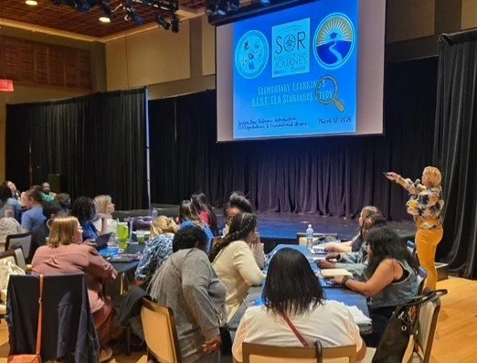 A woman giving a presentation to an audience at a conference or seminar in a large room with a stage and a screen displaying a slide about collaborative learning.