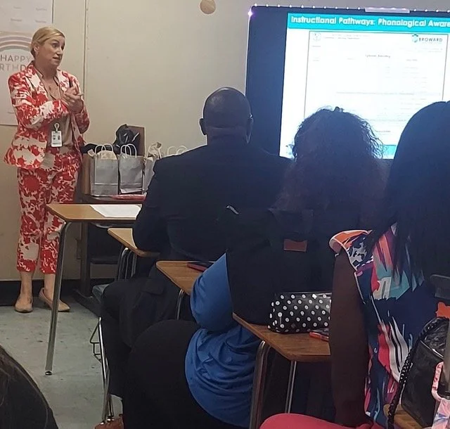 A woman in a red and white patterned dress standing in front of a classroom giving a presentation, with a large screen displaying a slide titled 'Instructional Pathway: Phonological Awareness', and three seated people facing her.