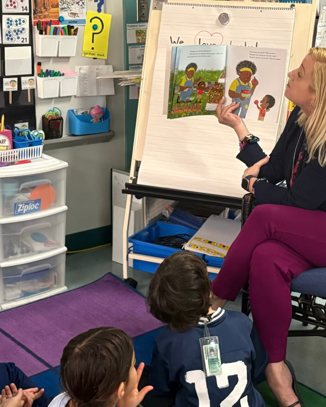 A woman reading a picture book to a group of children seated on the floor in a classroom.