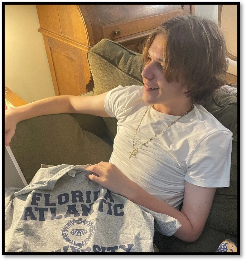 A young man with shoulder-length brown hair, wearing a white t-shirt and a gold chain necklace, is sitting on a green couch smiling. He is holding a gray Florida Atlantic University T-shirt.