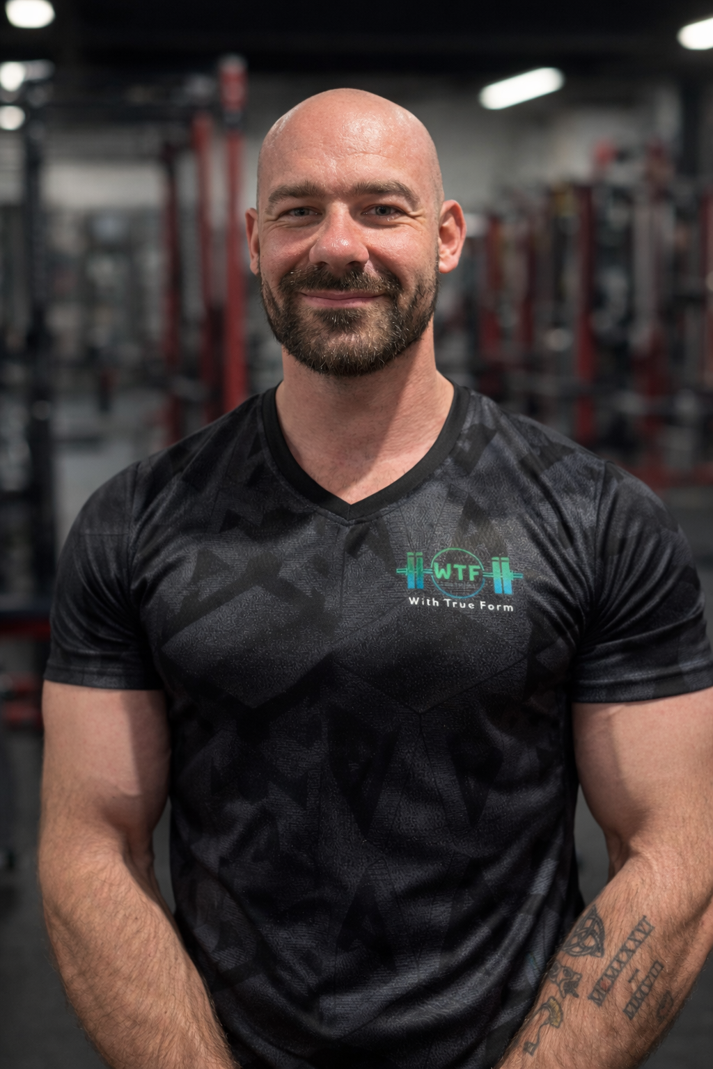 A muscular man with a shaved head and beard in a gym, smiling and crossing his arms - this is the WTF coach, Liam.