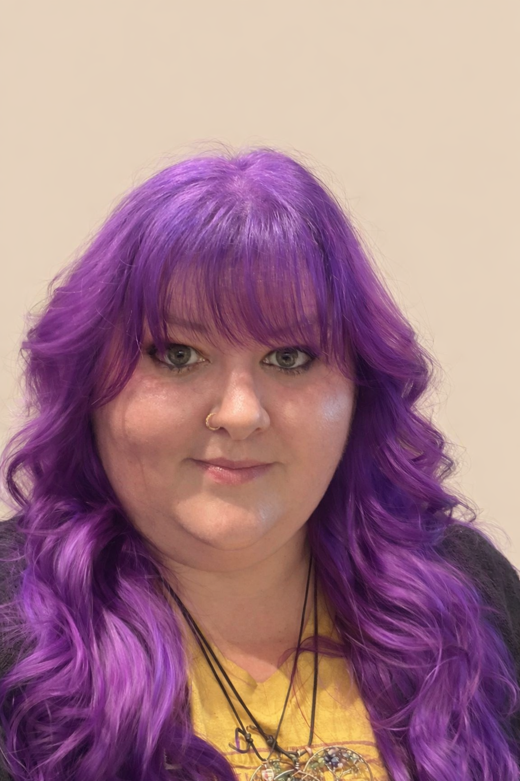 A woman with long, wavy, purple hair and bangs, wearing a yellow top, black cardigan, and layered necklaces, standing against a plain light-colored background.