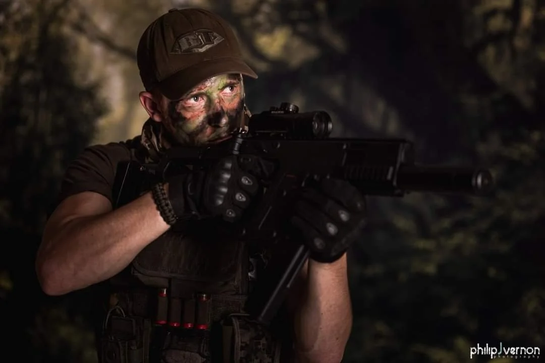 Soldier with camouflage face paint aiming a scoped rifle, wearing tactical gear and a cap, in a dark wooded environment.