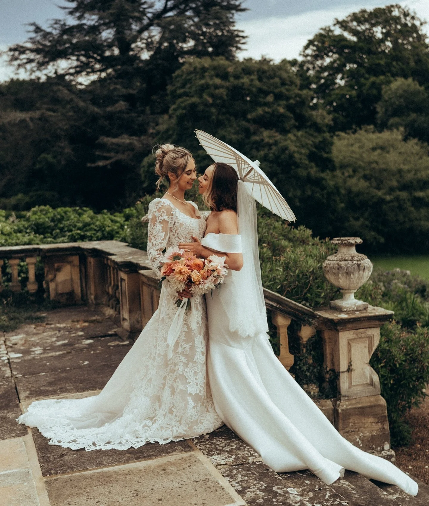 Two women in wedding dresses sharing a moment on a stone terrace, with greenery and trees in the background. One woman holds a bouquet of peach-colored flowers, and the other holds a lace parasol.
