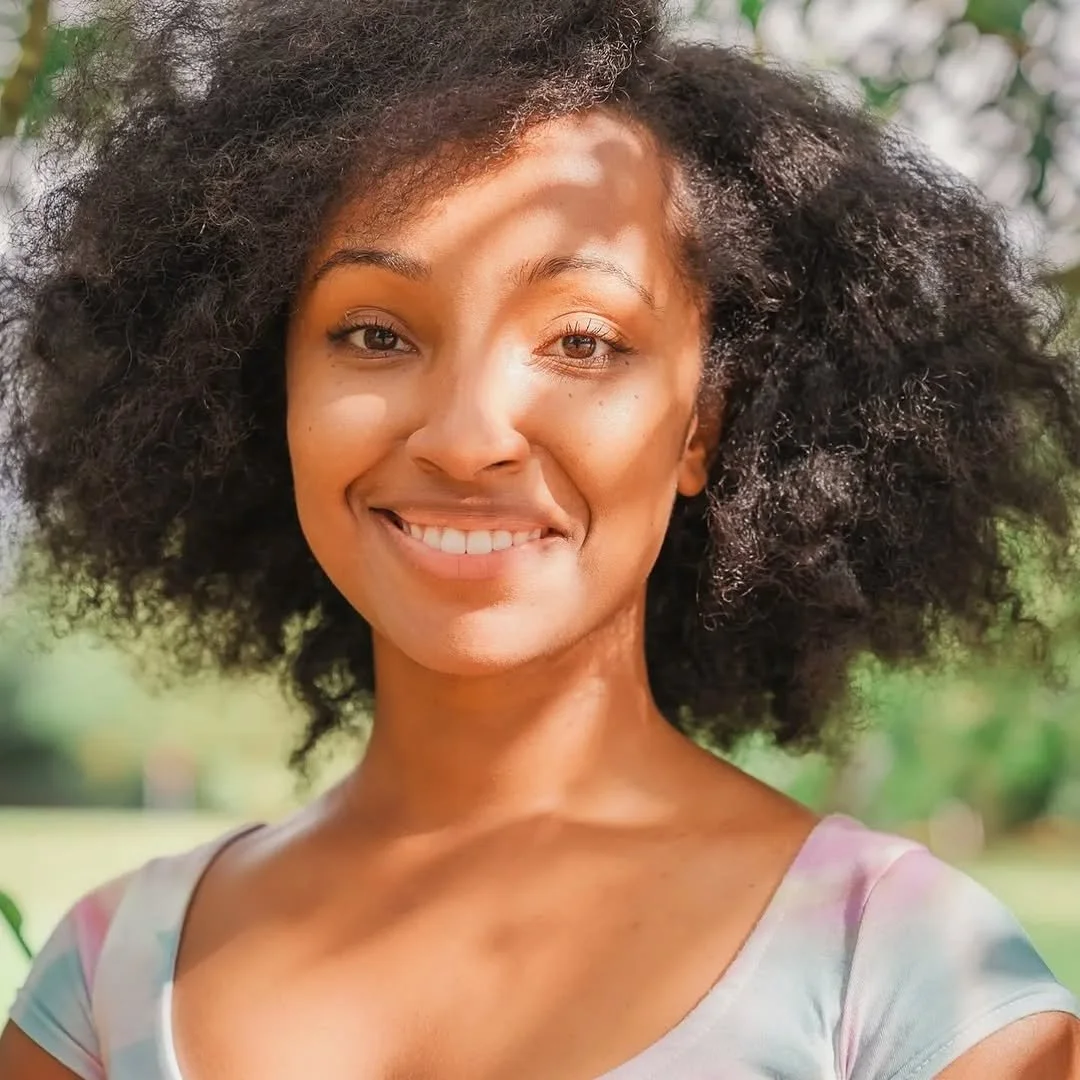 A smiling woman outdoors with curly black hair, wearing a colorful top, in a natural setting with greenery in the background.