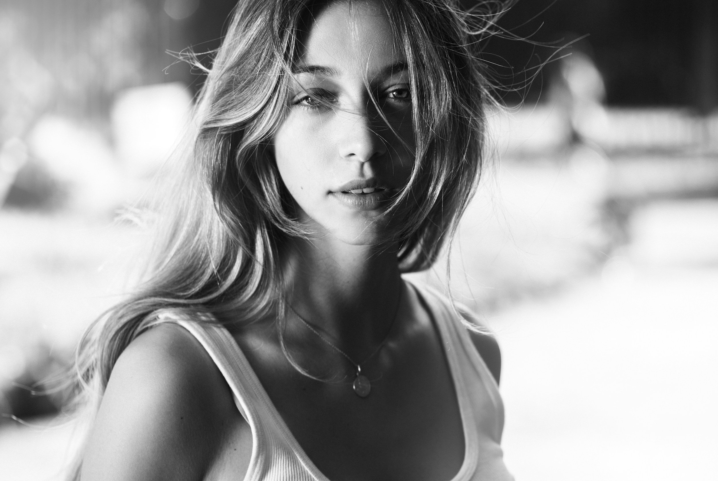 A black and white close-up portrait of a young woman with long, wavy hair. She is looking into the camera with a slightly parted mouth, wearing a tank top and a necklace with a small pendant.