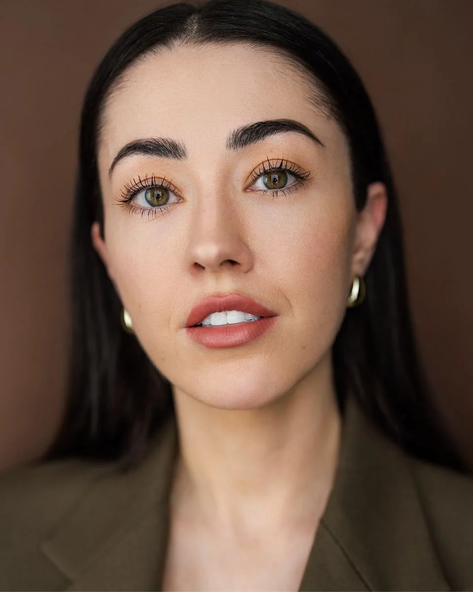 A close-up portrait of a woman with dark hair, hazel eyes, and natural makeup, wearing a brown blazer and gold hoop earrings, against a brown background.
